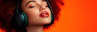 Young African American woman with red curly hair wearing wireless headphones and bright red lipstick against vibrant orange background, eyes closed in musical enjoyment.