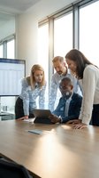 Diverse business team of four professionals gathered around tablet reviewing data in modern office with bright windows and natural light during collaborative meeting.