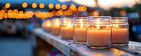 Glowing candles in glass jars lined up outdoors at dusk, with soft bokeh lights creating atmospheric mood for romantic or memorial events.