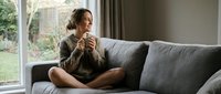 Young woman relaxing on gray sofa with hot beverage, enjoying peaceful morning moment in cozy living room with natural window light for lifestyle content.