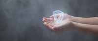 Cupped hands holding glowing transparent house model with rainbow light effects against dark smoky background for real estate and home protection concepts.