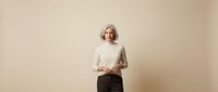Middle-aged Caucasian businesswoman in cream sweater and dark pants standing confidently against neutral beige background for corporate presentations.