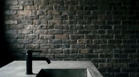Matte black modern faucet on white marble countertop against dark brick wall backdrop. Contemporary bathroom or kitchen design element for interior styling and home renovation projects.