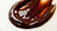 Glossy dark brown liquid substance with swirled texture and golden highlights on cream background, suitable for cosmetics, skincare, and beauty product marketing materials.