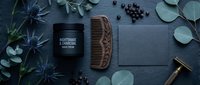 Men's grooming essentials with wooden beard comb, moisturizer jar and razor on dark wooden background with eucalyptus leaves and blue thistles.