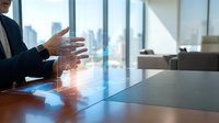 Professional businessman gesturing during presentation in modern office conference room with city skyline view through large windows.