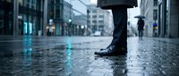 Professional businessman walking on wet city street pavement in formal black leather shoes during rainy weather with urban office buildings background.