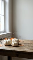 White pumpkins and autumn leaves on rustic wooden table near window creating cozy fall seasonal home decor arrangement for harvest celebration.