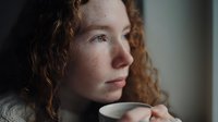 Young woman with curly red hair and freckles gazing thoughtfully while holding a cup of coffee in soft window light.