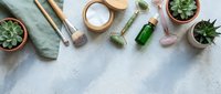Flat lay arrangement of skincare and wellness products with succulent plants, green glass bottles, wooden brushes, and cosmetic jars on textured surface for beauty and spa content.