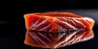 Fresh raw salmon fillet piece with glossy texture on black reflective surface, close-up view showing rich orange-red color and marbled pattern in dramatic lighting.