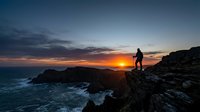 Silhouette of hiker standing on rocky cliff overlooking ocean at sunset, golden sky reflecting on water, dramatic coastal landscape for travel and adventure themes.