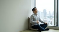 Young Asian man in casual sweater sitting cross-legged by large window overlooking city skyline, peaceful meditation pose in modern minimalist interior space.