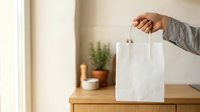 Hand holding white paper bag with handles on wooden table with potted plant and home decor in background, suitable for retail, shopping, and packaging design projects.