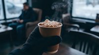 Hand in black glove holding steaming hot chocolate with marshmallows in paper cup at cozy winter cafe interior.
