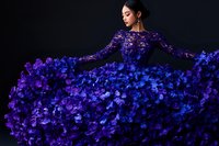 Young Asian woman in luxurious purple floral evening gown with long sleeves poses gracefully against dark background, dress flowing with dimensional flower details.