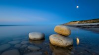 Tranquil seascape with round boulders on calm water reflecting moonlight, coastal cliffs and blue twilight sky creating serene natural composition.
