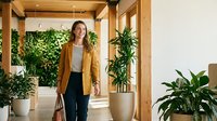 Professional young Caucasian businesswoman in mustard blazer walking through modern office lobby with lush green plants and wooden architectural elements.
