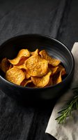 Crispy golden sweet potato chips seasoned with herbs in black ceramic bowl on dark textile background for healthy snacking and gourmet cooking concepts.