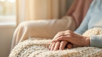 Elderly woman's hands resting peacefully on knitted blanket in warm sunlit room, conveying comfort, aging gracefully, and serene home atmosphere.