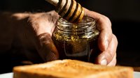 Beekeeper holding jar of golden honey with wooden dipper over fresh honeycomb, dripping liquid sweetener for food production and natural ingredient use.