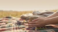 Hands holding warm coffee mug on colorful blanket during outdoor picnic in golden sunlight creating cozy autumn camping atmosphere for relaxation and leisure.