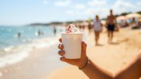 Cropped hand holding cold beverage cup with whipped cream topping at sandy beach with ocean waves and blurred vacationers in background, summer refreshment concept.