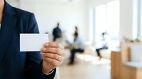 Professional in dark blazer holding blank white business card with blurred office background and colleagues, suitable for corporate identity, networking, and business branding materials.