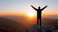 Male hiker celebrating success on mountain peak at sunrise with arms raised, silhouette against golden sky and valley landscape below.