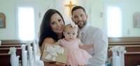 Young family with mother, father, and toddler daughter in pink dress posing together inside modern church with white walls and wooden pews during religious celebration or christening ceremony.