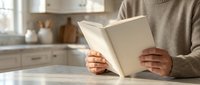 Adult person wearing beige sweater reading open book at kitchen table with blurred background, suitable for education, leisure, and lifestyle content.