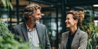 Mature businessman and businesswoman smiling at each other in modern green office space with plants, representing professional connection and workplace wellbeing.