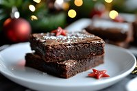 Rich chocolate brownies with festive sprinkles on white plate against blurred Christmas lights and decorations background, close-up view showing fudgy texture.