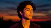 Young Hispanic man with curly hair meditating peacefully at sunset with eyes closed, warm golden hour lighting creating serene atmosphere for wellness content.