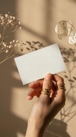 Female hand holding blank white business card with golden rings and dried flowers in warm sunlight shadows for branding mockup and stationery design.