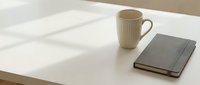 Minimalist workspace with cream ribbed mug and gray notebook on white desk, natural sunlight creating soft shadows, modern office setup for productivity and design projects.