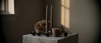 Minimalist holiday table setting with black taper candles, dried floral arrangements, and marble pedestal against neutral wall with natural window light.