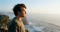 Young man with curly hair in knit sweater gazing at ocean waves from rocky cliff during golden hour sunset, contemplative mood for wellness and lifestyle content.