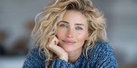 Blonde woman with blue eyes and wavy hair smiling gently while resting chin on hand, wearing cozy blue sweater against soft blurred background.