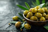Fresh green olives in wooden bowl with olive leaves on dark stone background, selective focus with copy space for Mediterranean cuisine and food design.