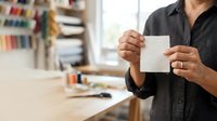 Male professional with medium skin tone holding blank white card in modern office workspace with bookshelves, suitable for business communication and corporate messaging concepts.