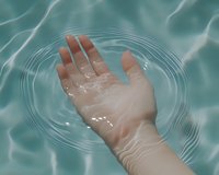 Human hand reaching into rippling turquoise water creating concentric circles, representing purity, meditation, and spiritual connection in wellness and spa settings.