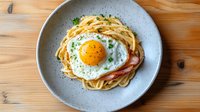 Spaghetti carbonara topped with sunny side up egg and crispy bacon on gray ceramic plate against wooden table background, garnished with fresh herbs.