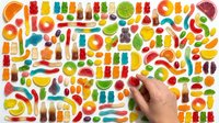 Colorful assortment of gummy candies, jellybeans, and sweet treats arranged in organized rows on white background. Hand reaching for candy selection.