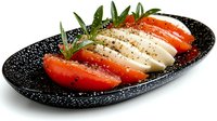Fresh mozzarella cheese slices with ripe tomato wedges and basil leaves on speckled black plate, drizzled with olive oil and black pepper seasoning.