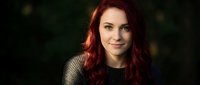 Young Caucasian woman with long red hair wearing gray sweater smiling at camera against dark background for portrait photography and lifestyle content.