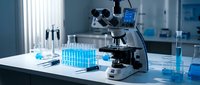 Modern laboratory microscope on white table with blue liquid test tubes, flasks, and scientific equipment in bright research lab.