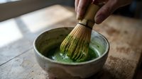 Hands whisking matcha powder in ceramic bowl with bamboo whisk, preparing traditional green tea ceremony beverage on wooden surface.