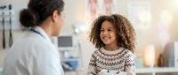 Young mixed-race girl with curly hair smiling during pediatric checkup with female doctor in modern medical office for healthcare consultation.