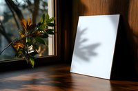 Blank white canvas mockup on wooden table near window with autumn leaves and natural shadows, minimalist home interior setting for artwork display.